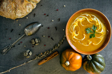Autumn menu. Top view photo of bowl with pumpkin soup, gem squashes, rosemary, pepper, fresh baked bread and silver spoon. Dark grey textured background. Healthy eating concept. 
