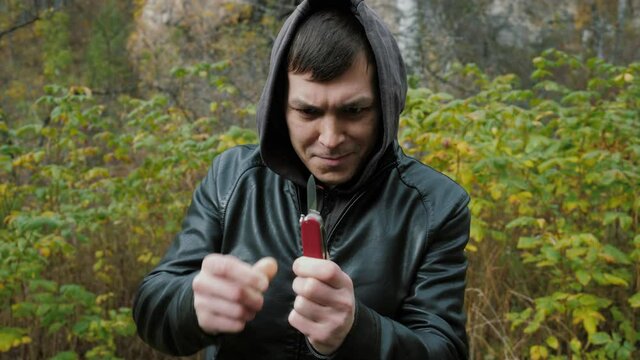 A surprised man in a hood tries to open a jackknife. A guy with dark hair in the forest deals with a knife. Medium shot.