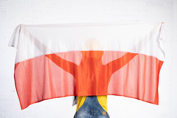 A construction worker stands back to the lens and holds the Polish national flag on her back. He has a yellow protective helmet on his head.
