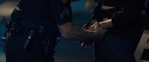 CLOSE UP Police officer handcuffs a suspect near police car, African-American Black criminal. Lights flashing in the background. Shot with anamorphic lens © supamotion