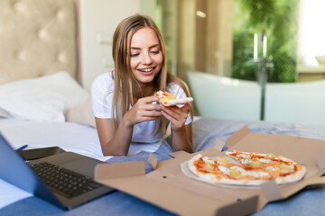 Young woman lying on a bed in bed at home while eating pizza and using laptop