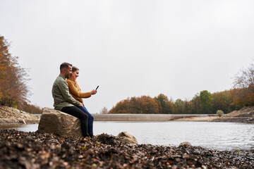 Wide angle of couple sitting in front of the lake looking at the mobile outdoors with a lake in the background. Copy space.