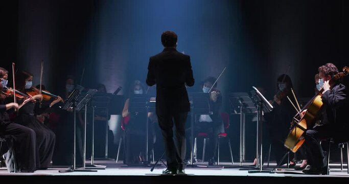 Cinematic shot of conductor directing symphony orchestra with performers with medical protective masks playing violins, cello and trumpet on classic theatre with curtain stage during music concert.
