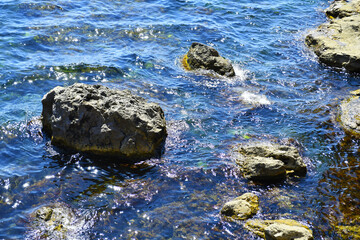 Multi-colored sun-drenched rocks and huge stones lying in the water near the shore. The rocky coastline of the Mediterranean Sea is washed by foamy waves.