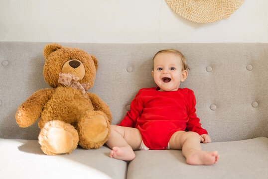 a baby girl in a red bodysuit sits on a gray sofa with a plush big bear and laughs