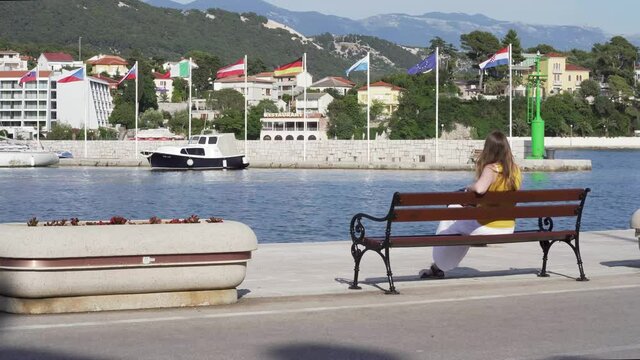 Girl in summer dress sitting on the bench next to sea with flags in back.