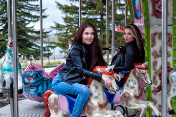 Fototapeta na wymiar Two young sexy women friends with long hair in leather jackets and jeans are sitting on an amusement ride in the park on an autumn day on a round carousel riding horses, posing and smiling.
