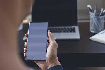 Mockup of man using and looking at blank screen mobile phone while working on laptop computer in office