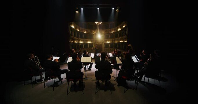 Cinematic shot of conductor directing symphony orchestra with performers playing violins, cello and trumpet on classic theatre with curtain stage during music concert with dramatic lights.