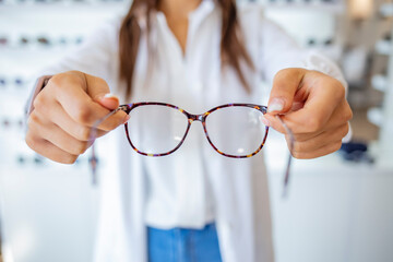Female optometrist, optician is standing with many glasses in background in optical shop. Stand with spectacles. Eyesight correction. Woman holds spectacles in hands. Closeup.