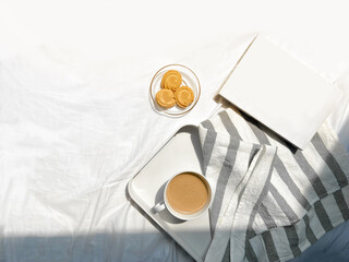 Hot coffee cappuccino in white cup in tray and strip fabric on bedsheet with cookies and book in Concept lazy weekend holiday morning breakfast with light from sunrise with warm drink for winter
