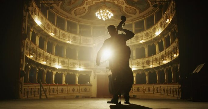 Cinematic silhouette of male cellist is playing cello on a classic theatre stage with dramatic lighting before start of a show.