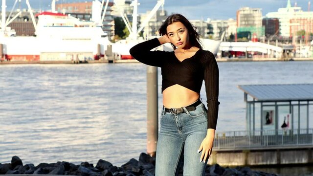 Eine junge sch&ouml;ne Frau mit langen schwarzen Haaren in einem schwarzen Cropped Top und Jeans posiert vor der Kamera in der Stadt. 
