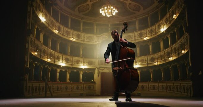 Cinematic shot of male cellist is playing cello solo on a classic theatre stage with dramatic lighting before start of a show.