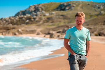 Handsome young blond man walks along the Black sea on the beach in sunny day