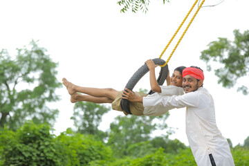 Cute indian child playing with his father on swing made by tyre and rope on tree at green field