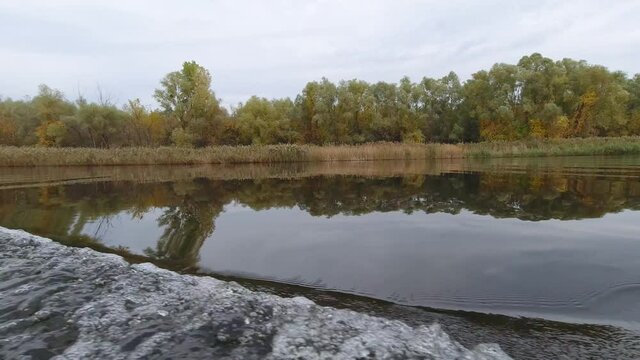 Reflection of the autumn forest in the water while the yacht moves along the river. Cloudy autumn day. Low point of view