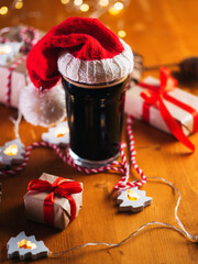 A beautiful glass of dark stout beer or ale on the Christmas table in a Santa Claus hat, wrapped gifts in Kraft paper
