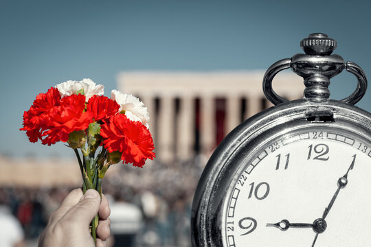 Hand holding red and white cloves next to big pocket watch in front of blurry Anitkabir mausoleum in November 10.  Old pocket watch showing time 9:05 AM, hour of the death of Ataturk.