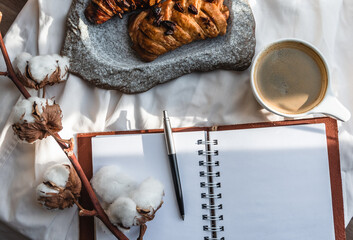 Fototapeta premium Coffee mug with croissant and empty notebook and pencil for business plan and design ideas onon white cloth from above, cozy and tasty breakfast, vintage toned