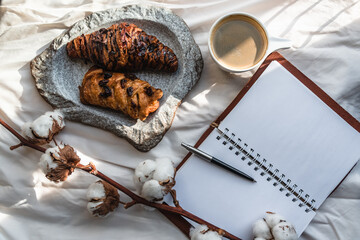 Coffee mug with croissant and empty notebook and pencil for business plan and design ideas onon white cloth from above, cozy and tasty breakfast, vintage toned