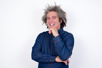 Handsome mature caucasian man with afro grey hair standing over isolated white backgrouOptimistic keeps hands partly crossed and hand under chin, looks at camera with pleasure. Happy emotions concept.