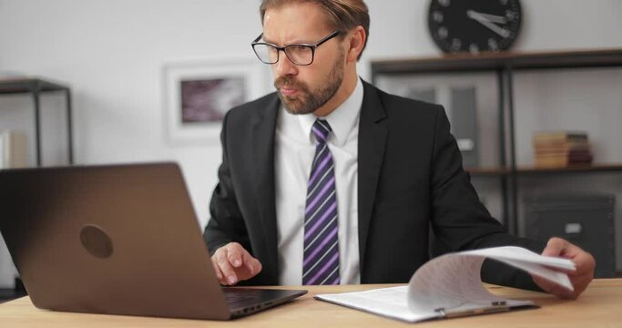 Confused bearded an in business suit and eyglasses rechecking all financial reports while sitting at modern office. Mature male using modern laptop for work.