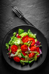 various fresh mix salad leaves with tomato, cucumber and red pepper on a black plate on Dark grey black slate background