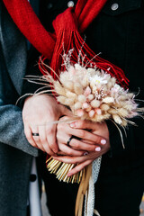 Black wedding rings on the bride and groom. The bride holds an autumn bouquet of dried flowers. a red scarf is tied around her neck. Image with selective focus and tinting