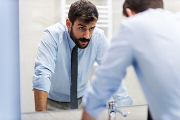 Young nervous attractive businessman leaning on the sink and looking himself in the mirror in restroom.