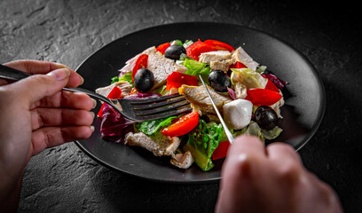 woman hands with knife and fork eat Fresh salad with chicken breast, cheese, black olives,red pepper, lettuce, fresh sald leaves and tomato on a black plate on Dark grey black slate background
