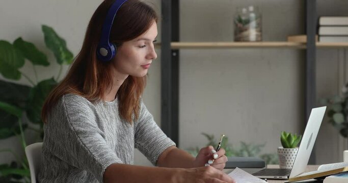 Focused young red-haired woman in headphones holding online video call meeting with tutor on laptop, discussing educational material, making notes, foreign language distant study e-learning concept.
