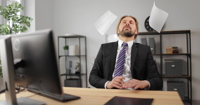 Bearded man in suit throwing papers around while sitting at table with modern computer. Mature man feeling tired from long office work.