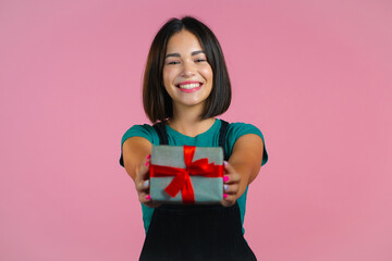 Excited woman holding gift box and gives it by hands to camera on pink wall background. Girl smiling, she is happy with present. Studio portrait