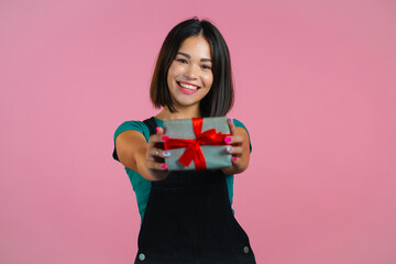 Fototapeta premium Excited woman holding gift box and gives it by hands to camera on pink wall background. Girl smiling, she is happy with present. Studio portrait