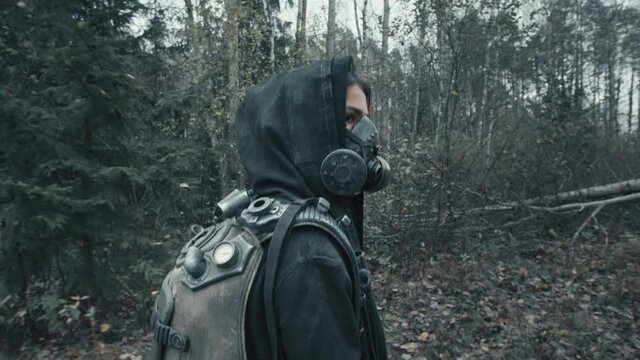 Following shot female wanderer in grunge mask hooded mantle walking in empty forest with bare trees. Apocalypse concept, survivor after war. Post apocalyptic world. Human of dystopian future