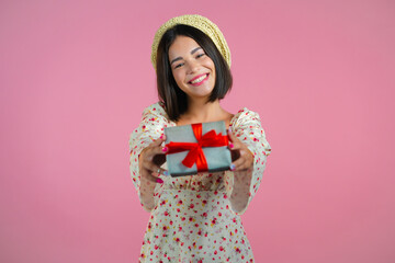 Excited woman holding gift box and gives it by hands to camera on pink wall background. Girl smiling, she is happy with present. Studio portrait