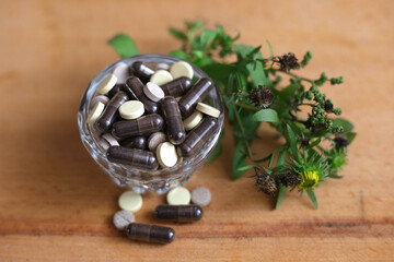 Herbal pills in glass cup on wooden background next to the herbs, top view. Natural medicines. Phyto capsules