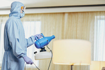 Man in blue protective suit holding disinfectant and standing in the room - obrazy, fototapety, plakaty