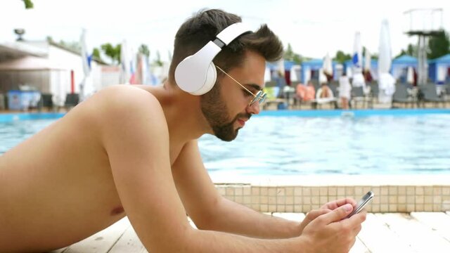 Handsome young man with mobile phone listening to music near swimming pool at resort