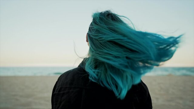 Hair toss of unique woman with teal blue or turquoise dyed hairstyle on sea beach background. Portrait of hipster girl with unusual hair enjoying summertime alone, demonstrate her fresh color.