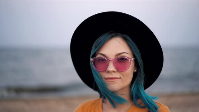 Portrait of hipster girl with unique fashionable blue hairstyle and hat on beach. Unusual woman in sunglasses with dyed colorful hair.