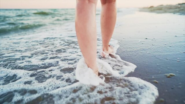 Legs of girl walking barefoot on wet sandy island beach. Beautiful feet of young woman near sea on sunset or sunrise. Slow motion.