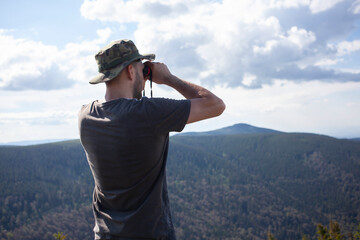 Man looking through binoculars wearing hat and tshirt at mountain hill nature forest landscape....