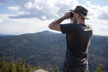 Man looking through binoculars wearing hat and tshirt at mountain hill nature forest landscape....