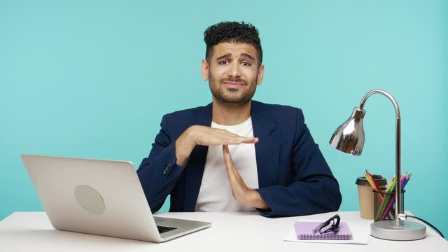 I need more time. Busy tired man in business suit showing timeout gesture, working at laptop and asking for break. Indoor studio shot isolated on blue background