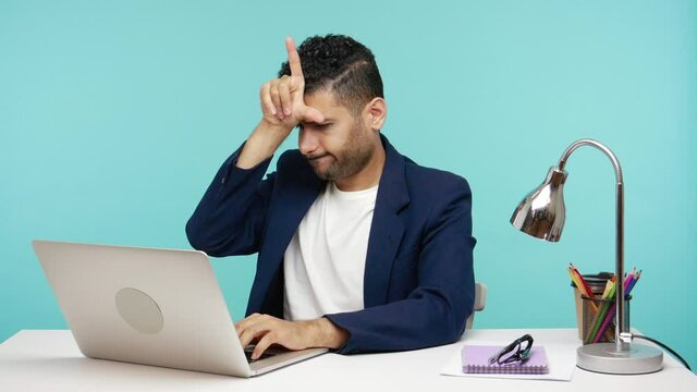 Depressed bearded man office worker showing loser gesture holding fingers near his forehead with sarcasm and negativity, failure at work. Indoor studio shot isolated on blue background