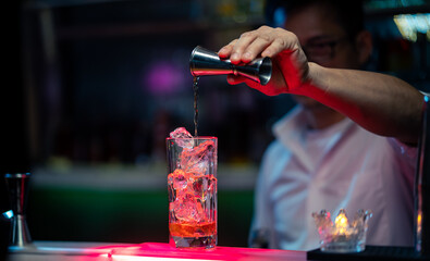 Professional Asian barman pouring liquor into long glass with ice cube on counter bar for serve in...
