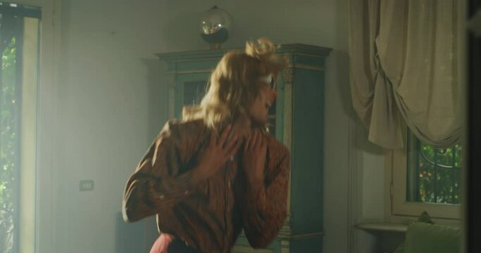Cinematic shot of young carefree happy smiling transgender woman wearing sunglasses, wig and fur coat is having fun to listening to the music, dancing crazy and jumping on sofa in living room at home.