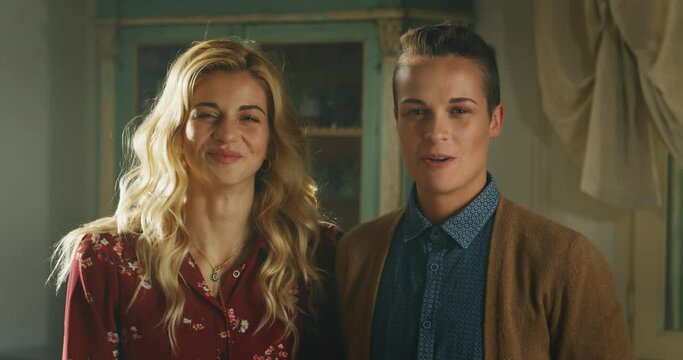 Cinematic shot of young happy homosexual female gay and transgender man couple is enjoying time together, kissing and smiling in camera at home. Concept of gender expression, identity and diversity.
